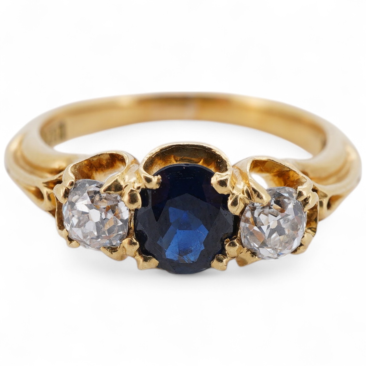 A Victorian sapphire and diamond three-stone ring, late 19th century, set with an oval sapphire between cushion-shaped diamonds totalling approximately 0.30 carats, to a gold mount of scrolled design, size J, maker's mar